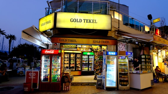 A brightly lit corner store named 'Gold Tekel' located on a busy street, featuring multiple refrigerators filled with beverages from brands like Coca-Cola, Pepsi, and Erikli. A vendor in the adjacent kiosk is serving a customer. There are visible advertisements and branding from Algida and other products. In the background, a few palm trees can be seen against a dusky sky.