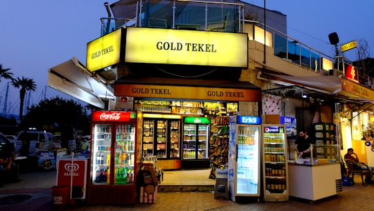 A brightly lit corner store named 'Gold Tekel' located on a busy street, featuring multiple refrigerators filled with beverages from brands like Coca-Cola, Pepsi, and Erikli. A vendor in the adjacent kiosk is serving a customer. There are visible advertisements and branding from Algida and other products. In the background, a few palm trees can be seen against a dusky sky.