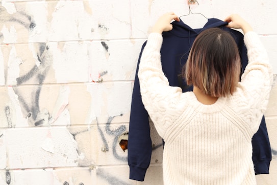 A person with short, dark hair is seen from behind, holding up a navy blue hoodie on a hanger in front of a textured wall. The wall has patches of peeling paint and graffiti, creating a rough and urban backdrop. The person is wearing a cream-colored knit sweater.