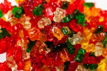 A large assortment of colorful gummy bears piled together, showcasing various translucent shades of red, green, yellow, and orange. The bears are tightly packed, with their glossy surfaces reflecting light, creating a vibrant and playful appearance.