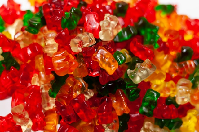 A large assortment of colorful gummy bears piled together, showcasing various translucent shades of red, green, yellow, and orange. The bears are tightly packed, with their glossy surfaces reflecting light, creating a vibrant and playful appearance.