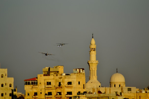 Two airplanes are flying in the sky above a cityscape featuring a prominent minaret and several beige-colored buildings. The lighting suggests either early morning or late afternoon.