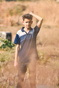 A young person stands outdoors in a natural setting, wearing a navy blue and white polo shirt with dark pants. Their hand rests on their head, and they are surrounded by blurred grasses and a soft-focus background of earthy tones.