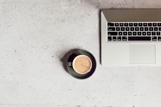 A modern laptop open on a wooden desk next to a steaming cup of coffee and a notepad.