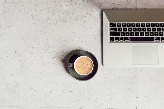 A laptop with an open keyboard sits on a light textured surface. Next to it, a cup of coffee in a dark saucer rests on the same surface.