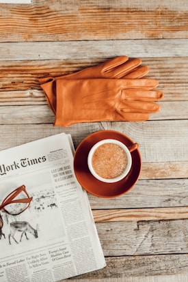 A cup of coffee in a brown saucer is placed on a rustic wooden table next to an open newspaper. A pair of orange leather gloves and brown-framed glasses rest on top of the newspaper.