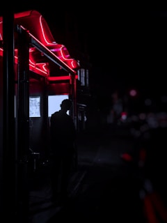 A shadowy figure standing beneath neon-lit arches, enveloped in deep purples and blacks, evoking a mysterious, cinematic vibe.
