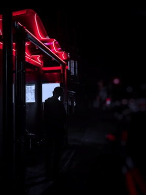 A shadowy figure standing beneath neon-lit arches, enveloped in deep purples and blacks, evoking a mysterious, cinematic vibe.