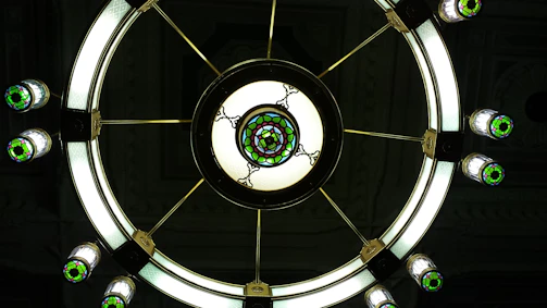 A large circular chandelier with stained glass decorations and illuminated bulbs. The design includes a central circular piece with intricate patterns and several smaller lights extending from the center, each featuring green and white stained glass.