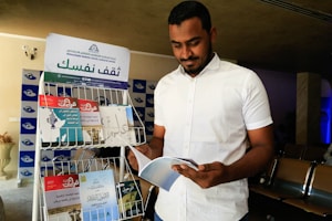 A man wearing a white shirt is standing indoors, reading a book. Next to him is a metal rack displaying various books and magazines, with an Arabic sign on top. The setting appears to be a cultural or educational event, as suggested by the material on the rack.