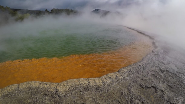 A geothermal pool with vivid colors, featuring a green central area surrounded by a bright orange rim. Dense mist rises from the water, enveloping the surrounding landscape. The rocky edge contrasts with the vibrant colors of the pool.