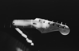 A black and white image focuses on the headstock of a Fender Stratocaster guitar, showcasing its tuning pegs and logo. The wooden texture is highlighted against a dark background, creating a classic and vintage aesthetic.