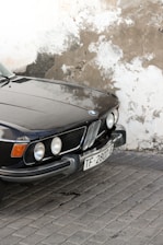 A vintage black car with a European license plate is parked on a cobblestone surface adjacent to a worn, textured wall. The car features a distinctive front grill and circular headlights, with a polished, reflective body.