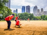 A baseball game taking place in a park, with players wearing red uniforms. The players are positioned around the field, framed by green trees and set against a backdrop of tall city skyscrapers. The field is sandy with clear skies overhead.