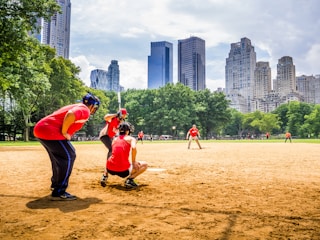 A baseball game taking place in a park, with players wearing red uniforms. The players are positioned around the field, framed by green trees and set against a backdrop of tall city skyscrapers. The field is sandy with clear skies overhead.