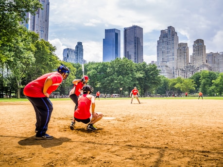 A baseball game taking place in a park, with players wearing red uniforms. The players are positioned around the field, framed by green trees and set against a backdrop of tall city skyscrapers. The field is sandy with clear skies overhead.