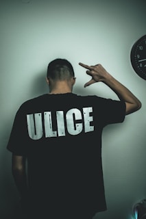 A person is standing with their back facing the camera, wearing a black T-shirt with the word 'ULICE' printed in bold white letters. The person's right hand is raised, making a gesture resembling a hand sign. A partially visible black clock is on the wall to the right.