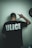 A person is standing with their back facing the camera, wearing a black T-shirt with the word 'ULICE' printed in bold white letters. The person's right hand is raised, making a gesture resembling a hand sign. A partially visible black clock is on the wall to the right.