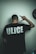 A person is standing with their back facing the camera, wearing a black T-shirt with the word 'ULICE' printed in bold white letters. The person's right hand is raised, making a gesture resembling a hand sign. A partially visible black clock is on the wall to the right.