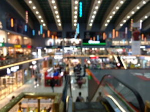 The image depicts the interior of a large shopping mall, with multiple shops, bright lighting, and people walking around. There are escalators and several levels visible, and the ceiling is adorned with decorative lights. The image is slightly blurred, making it difficult to identify specific details.