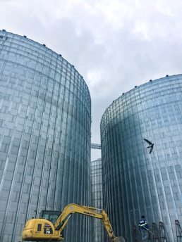 Two large industrial silos dominate the scene, constructed from metallic materials with a corrugated appearance. A yellow Komatsu excavator is visible in the lower foreground, along with a person who appears to be working near the base of one of the silos. A bird can be seen flying in the sky above.