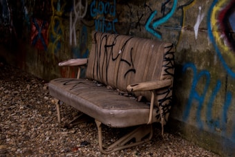 An old, worn-out sofa sits against a graffiti-covered wall. The fabric of the sofa is stained and torn, with various tags and markings drawn in black ink across its surface. The surrounding ground is covered in gravel, adding to the grungy and neglected atmosphere.