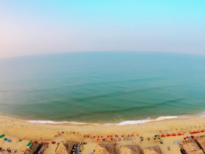 Aerial view of a vast beach with light golden sand and a calm, turquoise ocean stretching to the horizon. Various colorful parasols, beach chairs, and thatched huts are lined up along the shoreline, while a few people are scattered along the beach and swimming in the water.