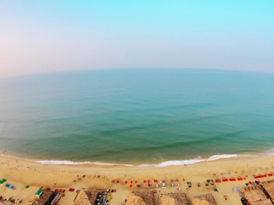 Aerial view of a vast beach with light golden sand and a calm, turquoise ocean stretching to the horizon. Various colorful parasols, beach chairs, and thatched huts are lined up along the shoreline, while a few people are scattered along the beach and swimming in the water.