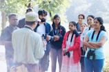 A group of people stand together outdoors, some wearing binoculars around their necks. They appear to be in a park or a natural setting, listening to a person in the foreground who seems to be speaking or guiding them. The background is lush with greenery and the mood is calm and educational.