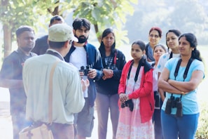 A group of people stand together outdoors, some wearing binoculars around their necks. They appear to be in a park or a natural setting, listening to a person in the foreground who seems to be speaking or guiding them. The background is lush with greenery and the mood is calm and educational.