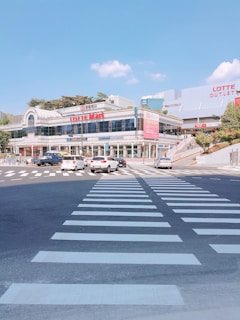 A wide pedestrian crossing leads to a modern shopping complex with large glass windows and visible signs. Several cars are driving along the road. The sky is clear with a few clouds, and the area appears busy but orderly.