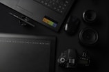 A flat lay of photography and design equipment, including a laptop, a graphic tablet, a pen, a digital camera, camera lenses, and a computer mouse. The items are arranged neatly on a dark surface, emphasizing a professional and creative workspace setting.