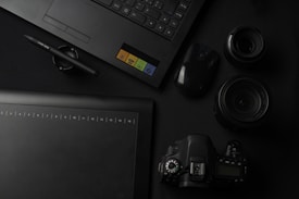 A flat lay of photography and design equipment, including a laptop, a graphic tablet, a pen, a digital camera, camera lenses, and a computer mouse. The items are arranged neatly on a dark surface, emphasizing a professional and creative workspace setting.
