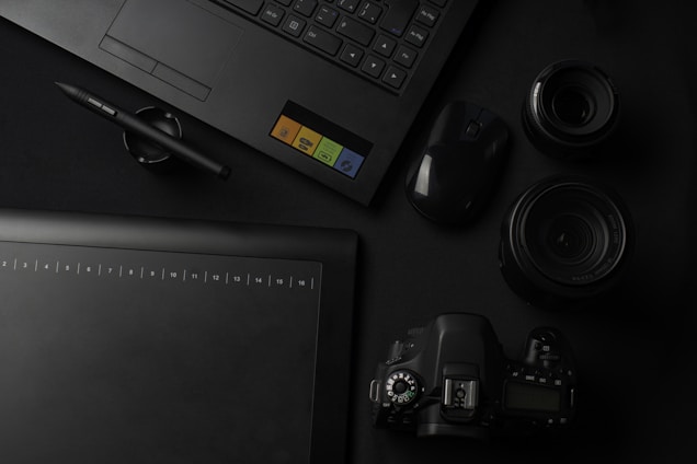 A flat lay of photography and design equipment, including a laptop, a graphic tablet, a pen, a digital camera, camera lenses, and a computer mouse. The items are arranged neatly on a dark surface, emphasizing a professional and creative workspace setting.