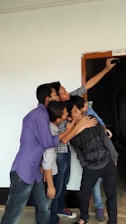 A group of five young people are gathered closely together, enthusiastically taking a selfie. They are standing near a doorway marked 'Class - X'. The hallway and walls around them have a simple, clean appearance.