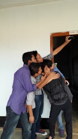 A group of five young people are gathered closely together, enthusiastically taking a selfie. They are standing near a doorway marked 'Class - X'. The hallway and walls around them have a simple, clean appearance.