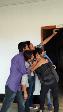 A group of five young people are gathered closely together, enthusiastically taking a selfie. They are standing near a doorway marked 'Class - X'. The hallway and walls around them have a simple, clean appearance.