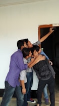 A group of five young people are gathered closely together, enthusiastically taking a selfie. They are standing near a doorway marked 'Class - X'. The hallway and walls around them have a simple, clean appearance.