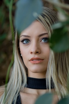 A person with long blonde hair and blue eyes is partially obscured by foliage, creating a sense of mystery and natural beauty. The person is wearing a black top and looking intently towards the camera.