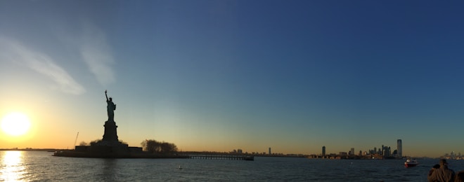 A panoramic view of a large statue on an island, with a setting sun casting a warm glow over the water. The skyline of a distant city is visible under a clear blue sky.