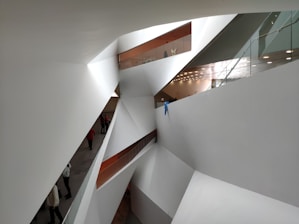 A modern architectural space with sharp, angular white walls and geometric shapes. The scene includes multiple levels connected by glass railings, and a few people walking on the lower level. The design features clean lines and a minimalistic style, with recessed lighting visible on one ceiling area.