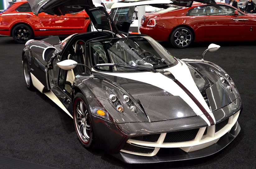 A sleek, modern sports car with a carbon fiber finish and white accents, displayed in an exhibition setting. The car features gull-wing doors open, showcasing its luxurious interior. In the background, there are two other high-end vehicles, one is red with a shiny exterior.