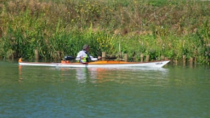 A person is paddling a kayak along a peaceful, green riverbank lined with tall grass and wildflowers. The kayak is white with orange accents, and the paddler is wearing a hat and life jacket.