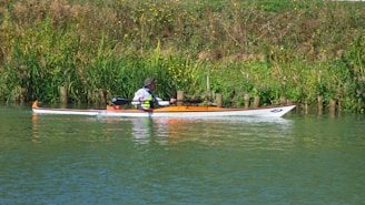 A person is paddling a kayak along a peaceful, green riverbank lined with tall grass and wildflowers. The kayak is white with orange accents, and the paddler is wearing a hat and life jacket.
