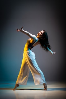 A female dancer is captured mid-movement in a dramatic pose, extending her arms outward with an expressive facial expression. She is dressed in a blue top and flowing white pants. The lighting casts a soft gradient on the background, transitioning from blue to a warm yellow, highlighting her silhouette.