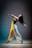 A female dancer is captured mid-movement in a dramatic pose, extending her arms outward with an expressive facial expression. She is dressed in a blue top and flowing white pants. The lighting casts a soft gradient on the background, transitioning from blue to a warm yellow, highlighting her silhouette.