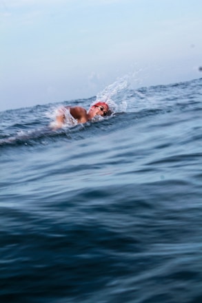 A person is swimming in open water, performing a freestyle stroke. The individual is wearing a red swim cap and swimming goggles. Water droplets are splashing around as the swimmer's arm moves through the water. The background is a vast, calm sea under a light blue sky with few clouds.