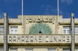 An old building featuring a decorative facade with a peacock design. The structure has large windows and visible weathering on the concrete. Above the windows, there is a sign with the words 'Novidade! Armazens Cunhas'. The building's design reflects vintage architectural styling.