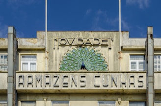 An old building featuring a decorative facade with a peacock design. The structure has large windows and visible weathering on the concrete. Above the windows, there is a sign with the words 'Novidade! Armazens Cunhas'. The building's design reflects vintage architectural styling.
