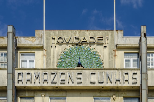 An old building featuring a decorative facade with a peacock design. The structure has large windows and visible weathering on the concrete. Above the windows, there is a sign with the words 'Novidade! Armazens Cunhas'. The building's design reflects vintage architectural styling.